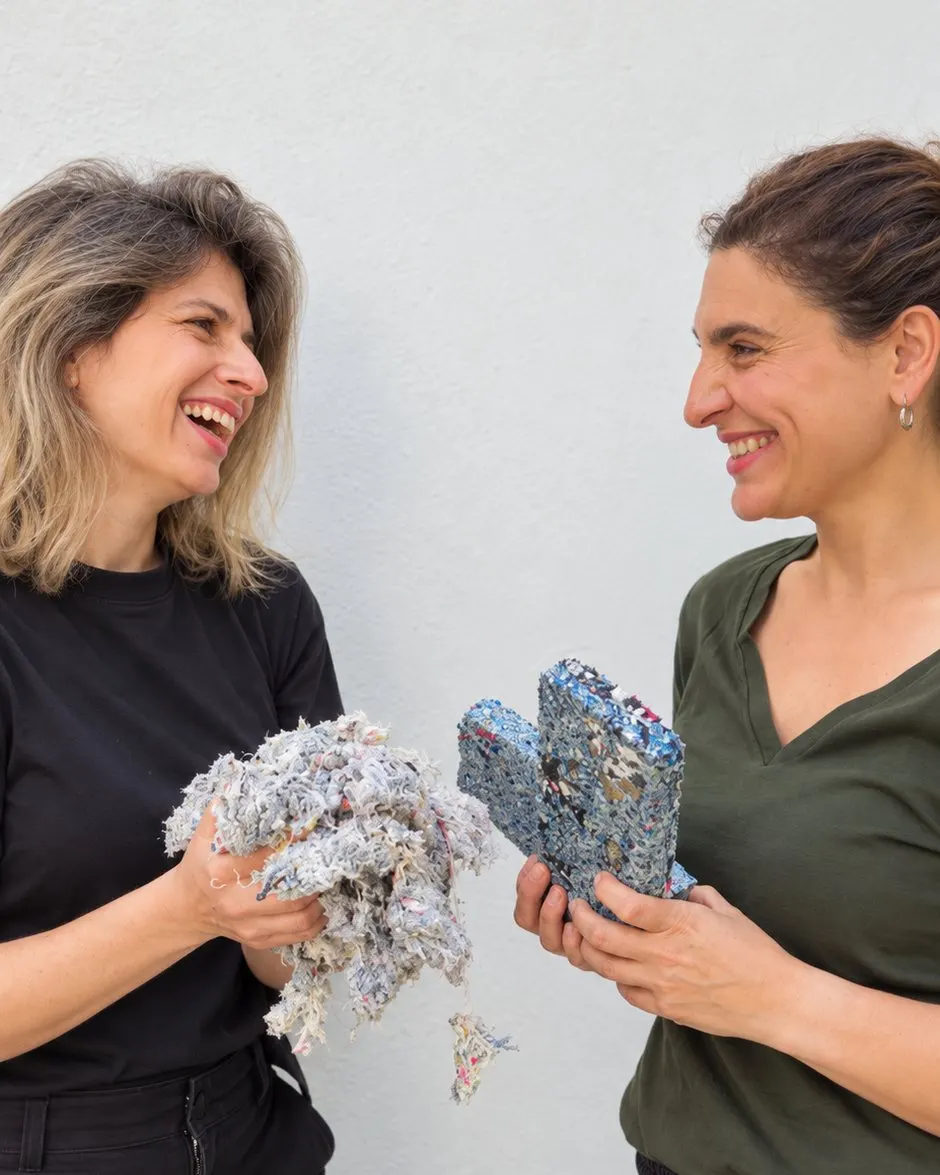 Verónica Litvinoff y Luciana Sabsay se miran y se ríen frente a una pared blanca: Verónica sostiene un puñado de recortes textiles y Luciana dos paneles de Requecho.