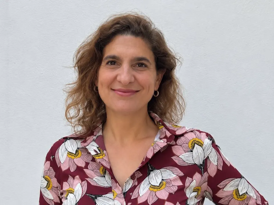 Retrato de Luciana Sabsay, sonriente, con camisa floral frente a una pared blanca.